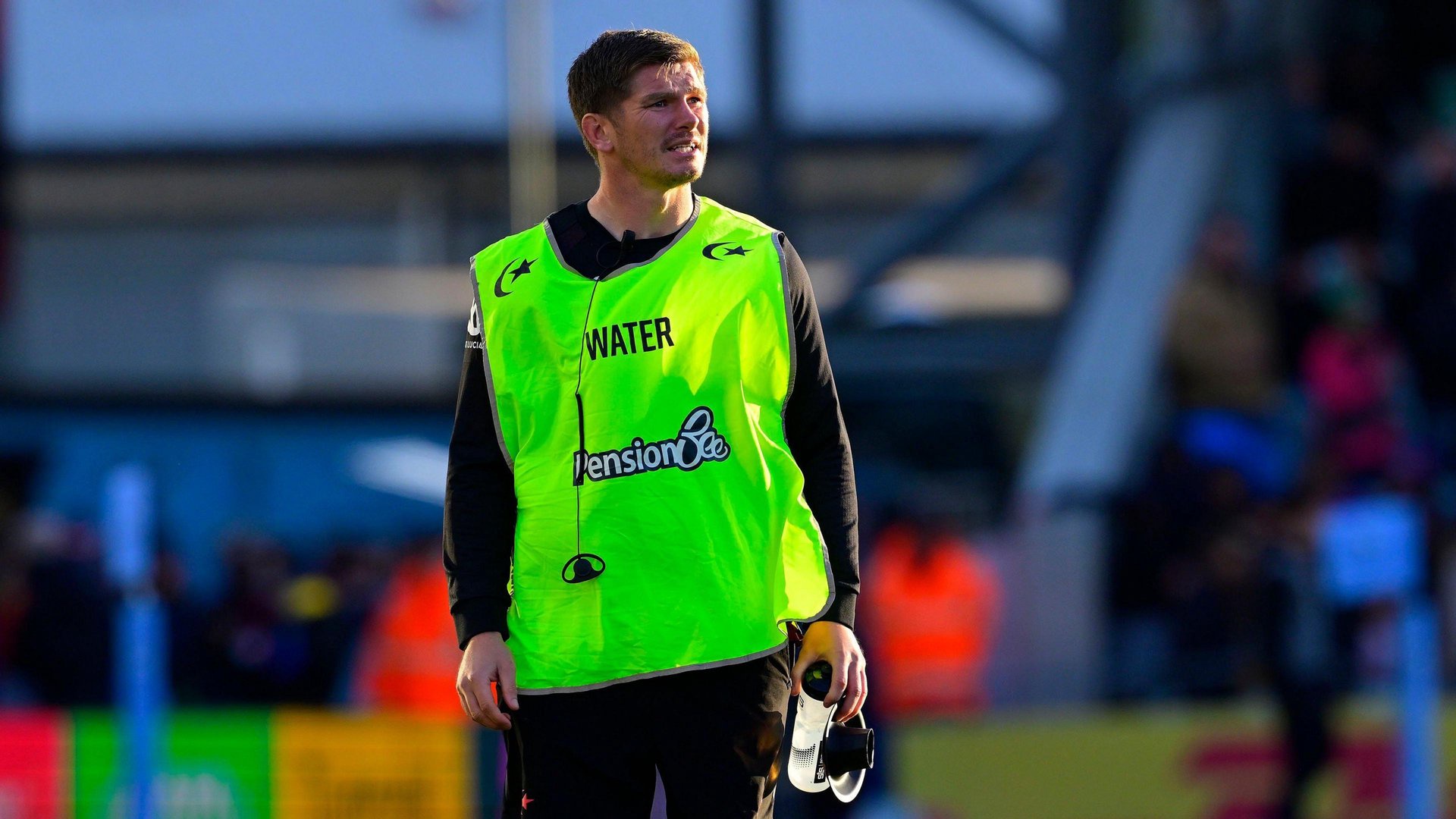 Owen Farrell of Saracens runs the water with a water bottle in his left hand while wearing a lime green fluorescent bib with the word "water" on it in capital letters.
