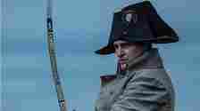 Joaquin Phoenix as Napoleon, wearing a military uniform and a bicorne hat, holding a sword