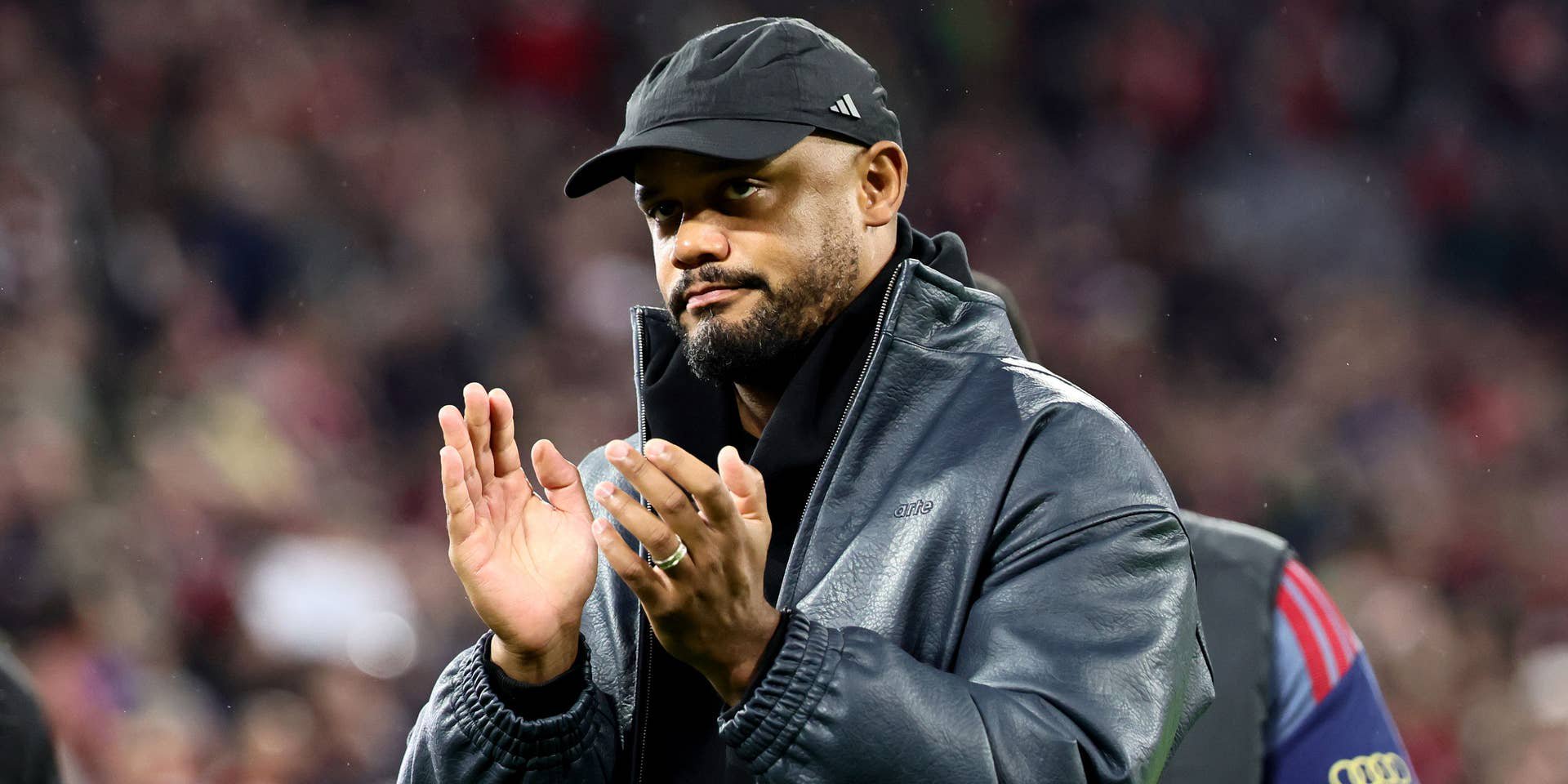 Ein Mann mit schwarzer Kappe und Jacke applaudiert in einem Stadion nach einem Fußballspiel.