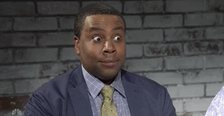 Kenan Thompson sitting in front of a brick wall, wearing a suit and tie, with a surprised facial expression