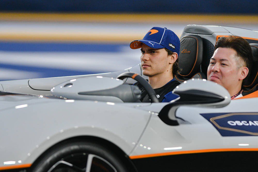 Driver in a blue McLaren cap in a silver open-top car with orange trim and "OSCAR" branding, at a McLaren event.