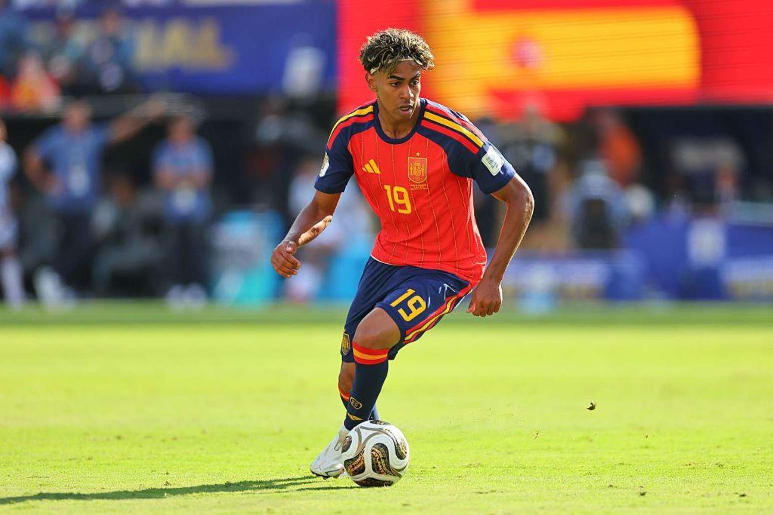 Lamine Yamal durante jogo da Espanha. Ele está chutando a bola e com a camisa vermelha da seleção.