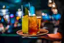 A server carries a tray with three drinks in a dimly lit bar: a green cocktail with a leaf, an orange-brown drink, and a glass of beer, with straws in the cocktails