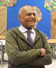 Algebra a civil right: Older Black man with gray hair in olive green cardigan, white shirt and brown tie, stands smiling widely with arms crossed in front of colorful bulletin board displays.