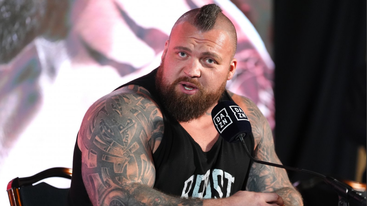 A heavily tattooed man speaks into a microphone during a press conference. He has a Mohican-style shaved head with a strip of hair down the middle and is wearing a black T-shirt.