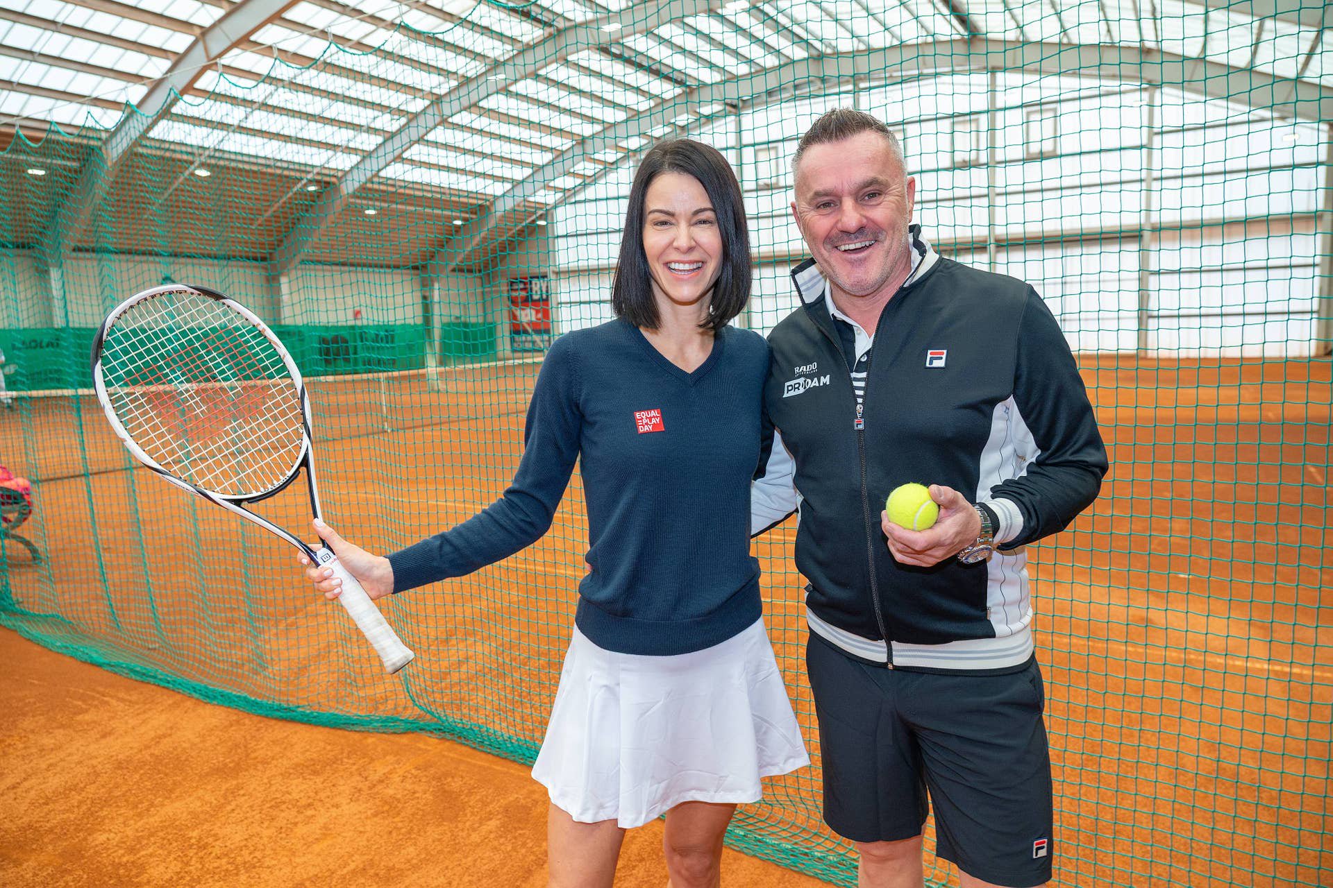 Zwei Personen stehen lachend auf einem Tennisplatz, eine hält einen Schläger, die andere einen Tennisball.