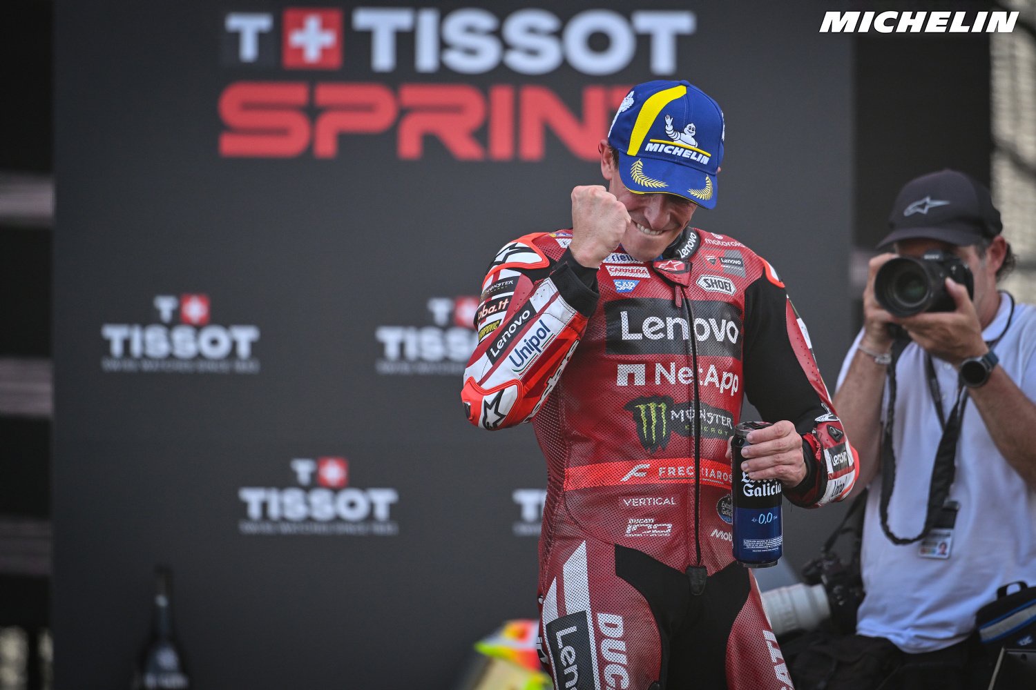 A MotoGP rider in red Ducati leathers with Lenovo and Monster logos, raising a fist on a Tissot podium stage, holding a Gaulois bottle.