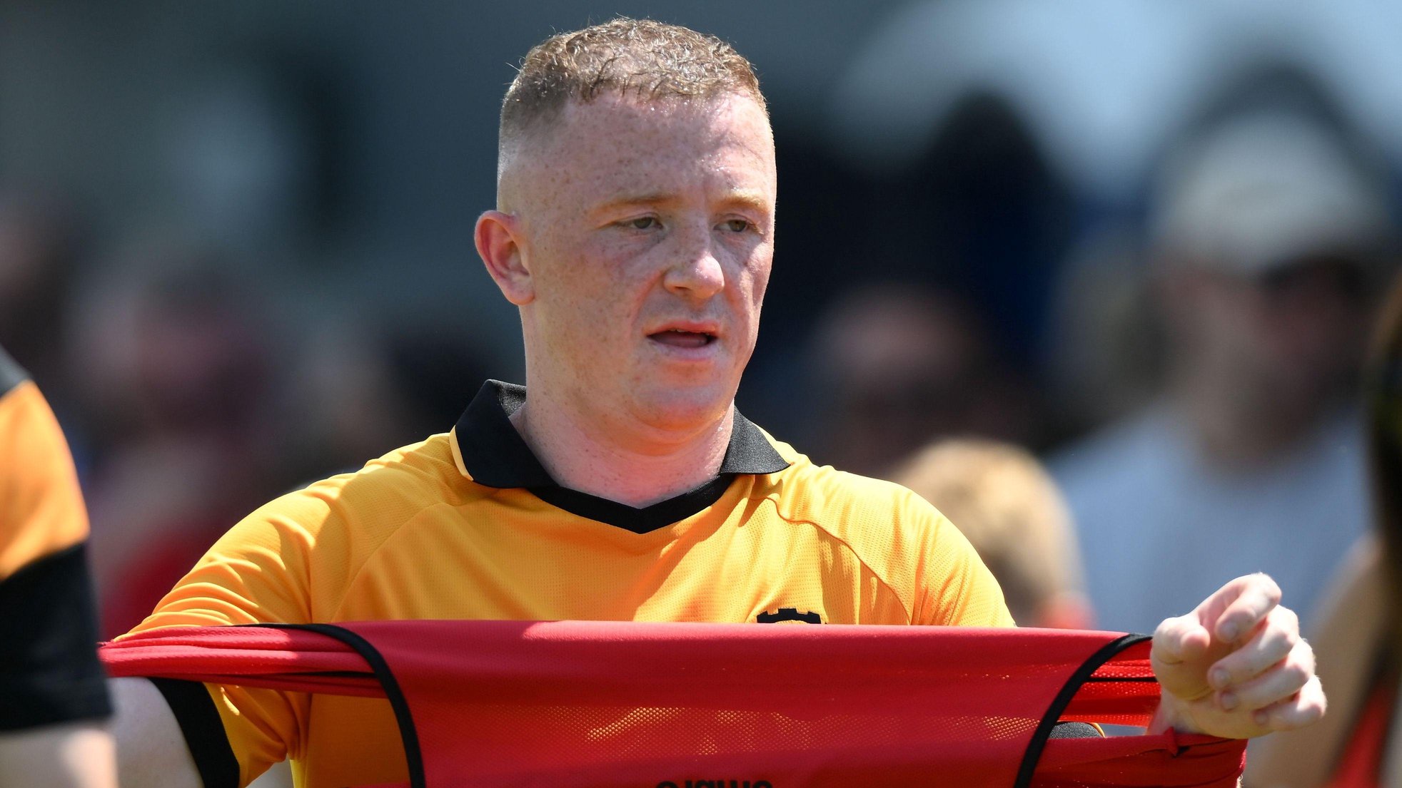 Cambridge United forward Shayne Lavery. He is wearing a yellow shirt with black collar and holding a red vest, which he is stretching out between his arms.