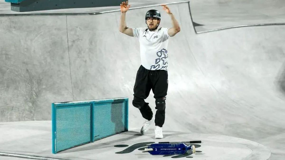 A man holding his arms up. He is wearing a helmet, a white T-shirt with black marks on it, black trousers and white trainers. He is in a scooter park, with a blue scooter lying on the ground to the right. There is a blue metal barrier next to him.