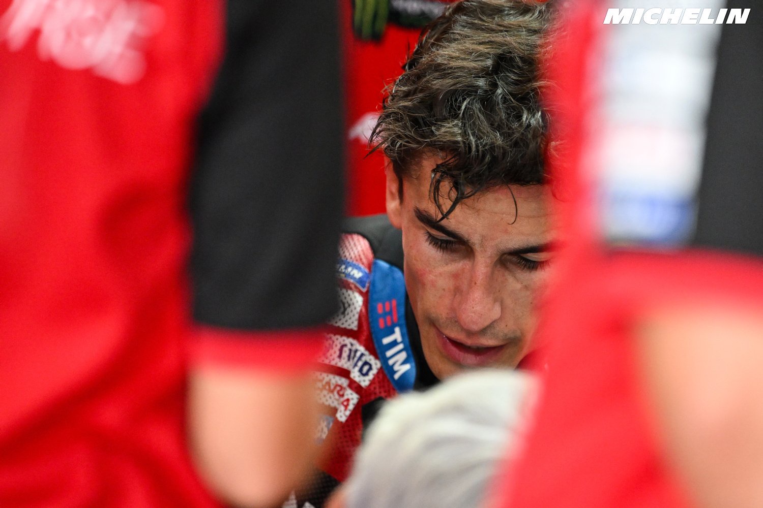 A rider in red team leathers with a TIM logo sits in the pit box, head bowed, surrounded by red-clad team members in a MotoGP paddock.