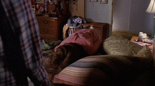 A person is seen lying facedown on a bed in a cluttered room with a dresser, clothes, and pillows visible. The setting suggests a relaxed, personal environment