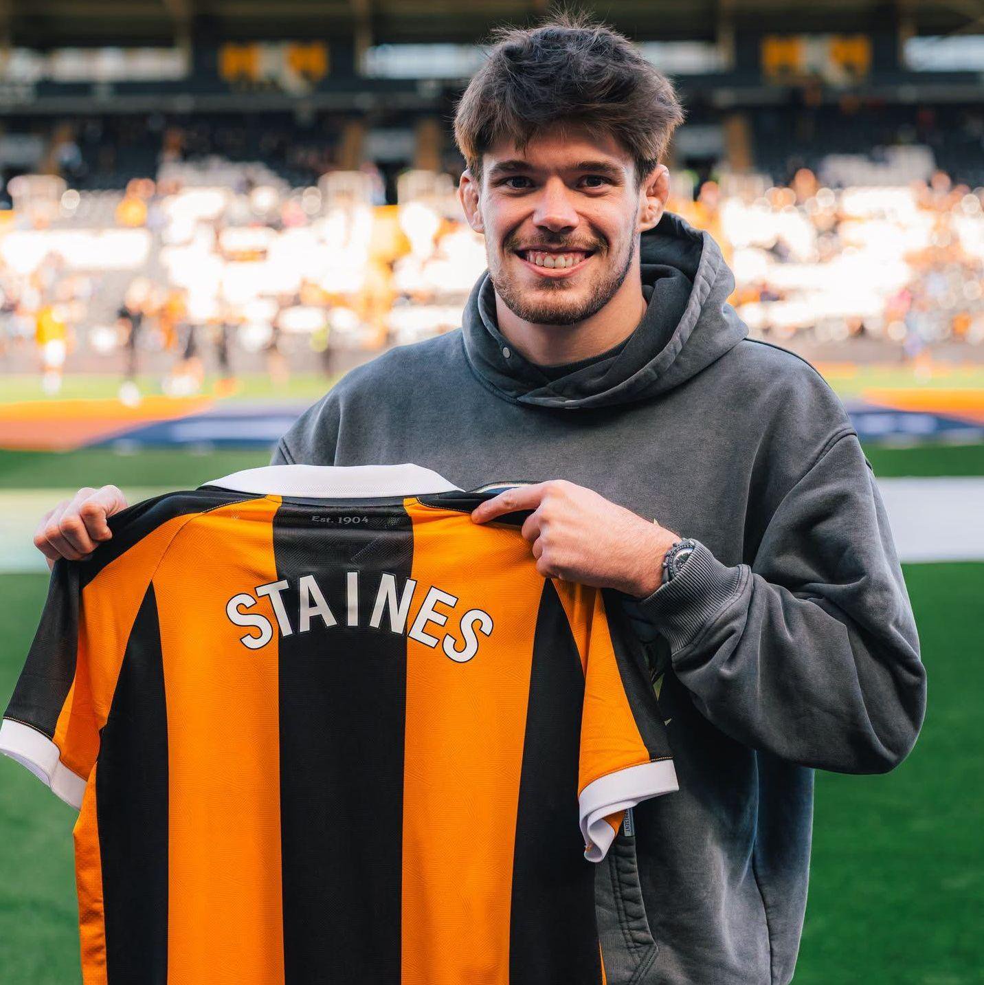 A man stands on a football pitch holding a black-and-amber striped Hull City shirt. The man is wearing a dark hooded sweatshirt and is smiling into the camera. Stadium seats and a green pitch are visible in the foreground.