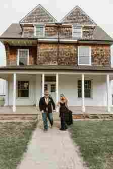 A couple walks hand in hand down a path in front of a rustic two-story house. The man is wearing a black suit with an orange tie and navy jeans, while the woman is dressed in a long, black strapless dress. Both are smiling as they stroll together.