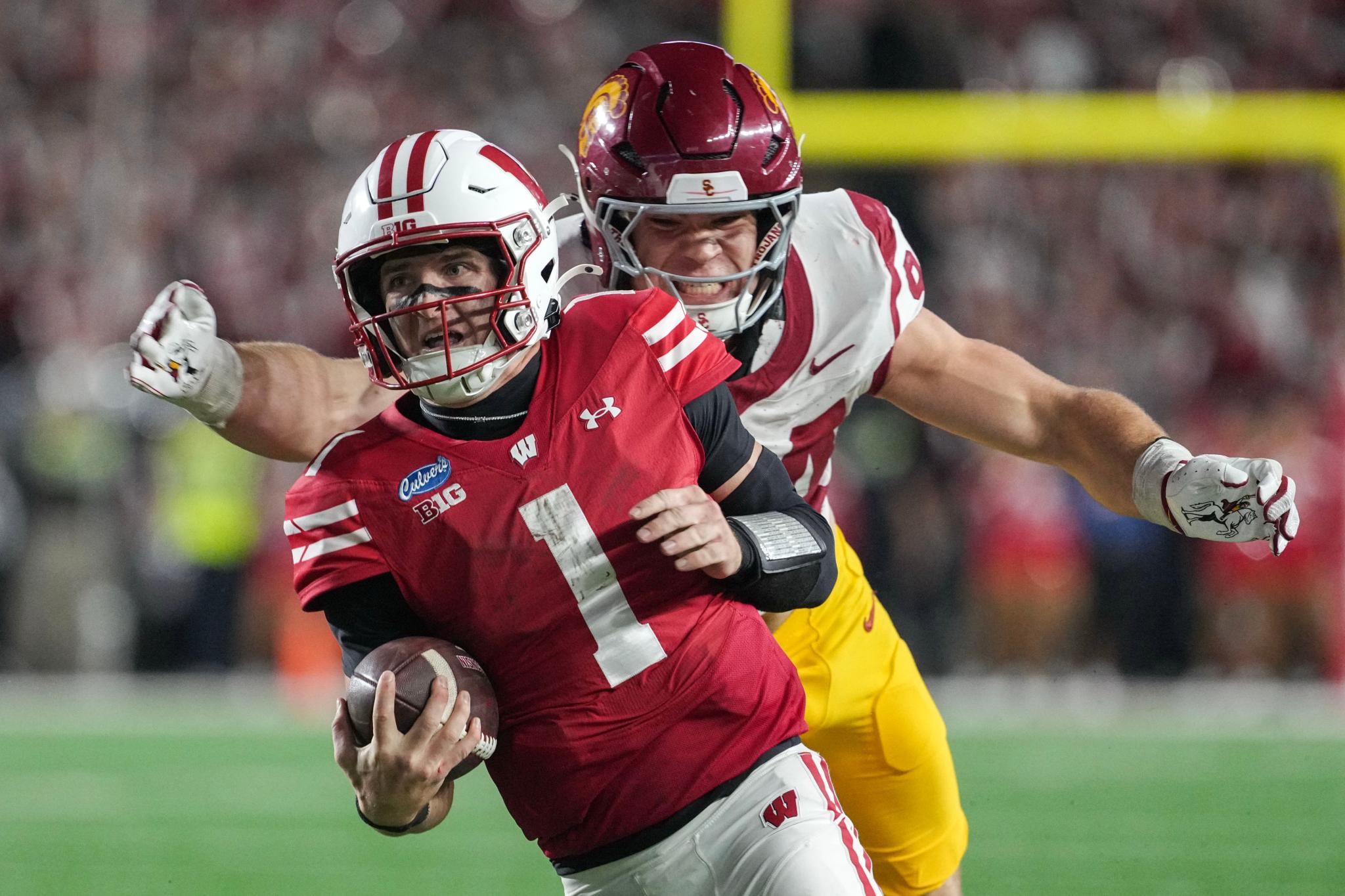 A Wisconsin football player in a red jersey runs with the ball as a USC player in a maroon and gold uniform pursues him.