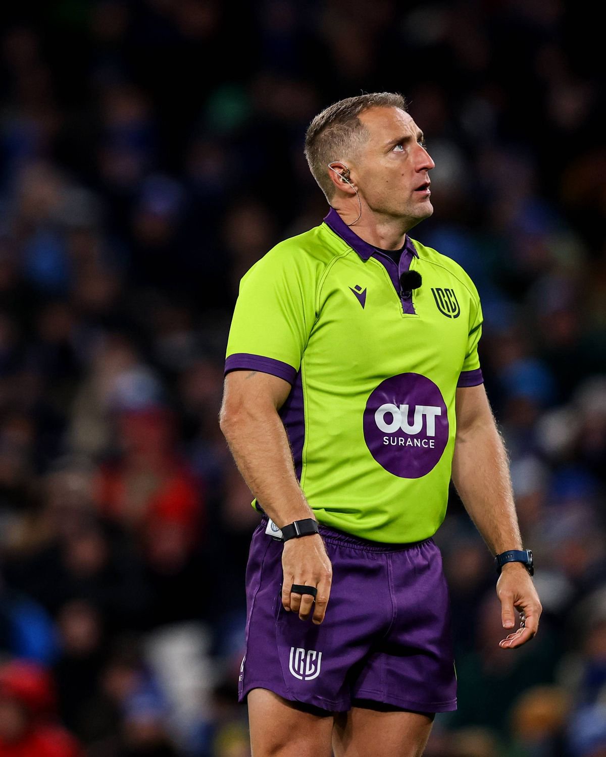 An individual, dressed in a neon green and purple sports uniform, stands on a field or court, facing a crowd. The person's uniform features the word "OUT" prominently on the front. The background is a blur of spectators, suggesting an ongoing event or game.