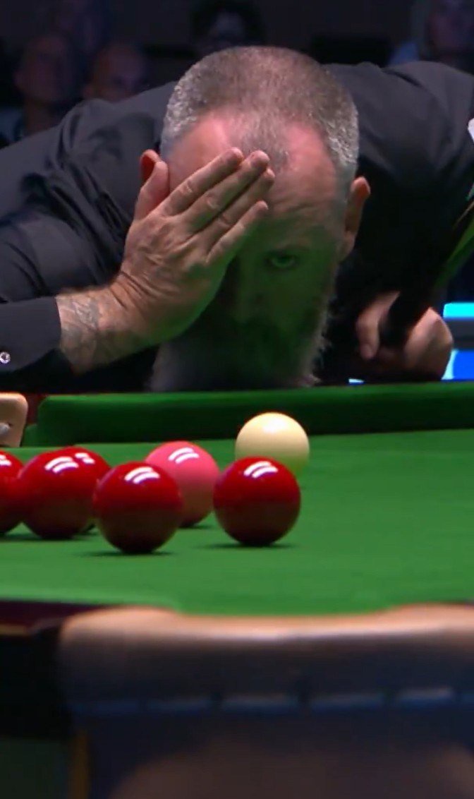 A snooker player bends over the table, placing his hand on his forehead to block the glare from overhead lights.