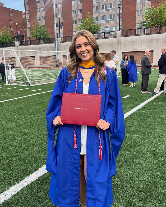 Young woman in blue graduation gown holding diploma on football field after ceremony
