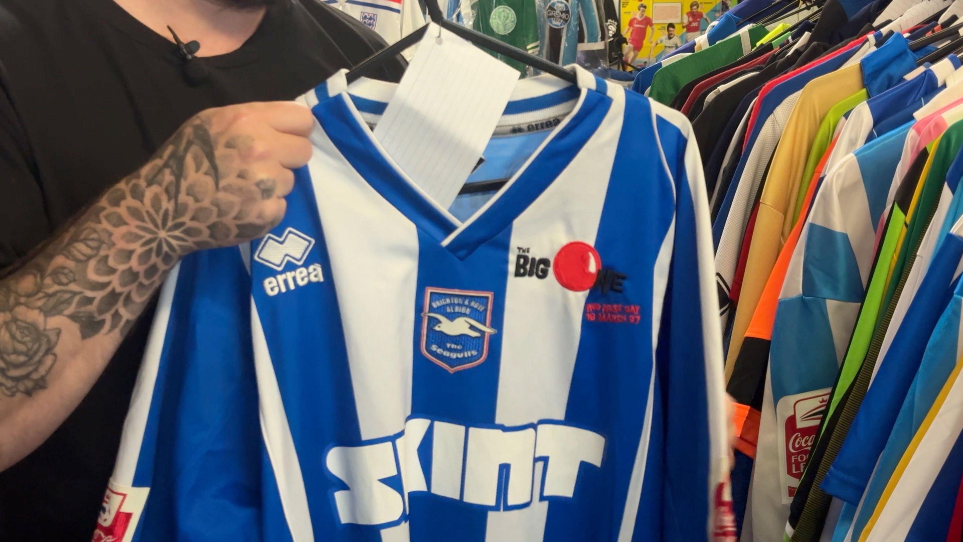 A man, wearing a black t-shirt, is holding up a blue and white striped football top with SKINT written on it in white. There is a badge with 'errea' written on it in white at the top left, with a red nose printed on the top left. To the right, dozens of football shirts are hanging.