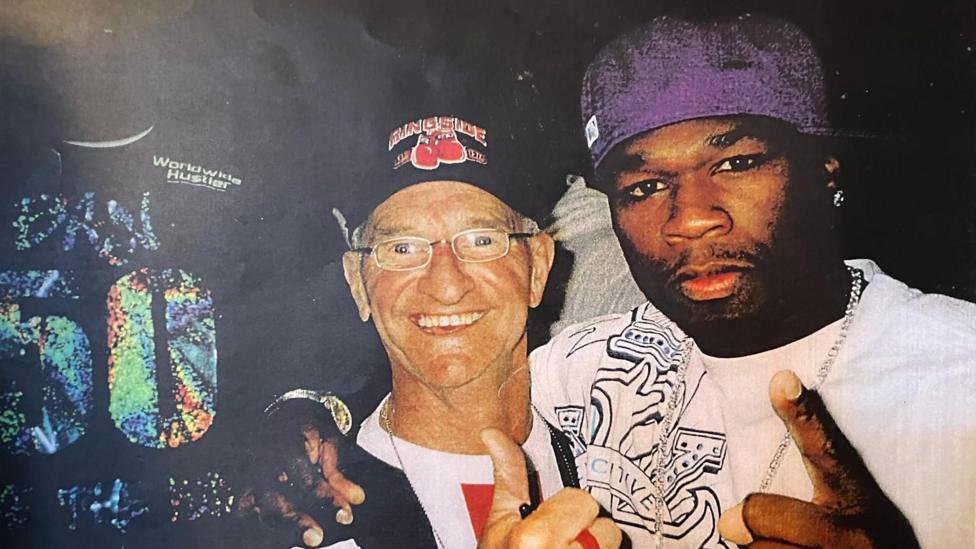 A man in his 60s standing next to 50 Cent, with their arms around each other.