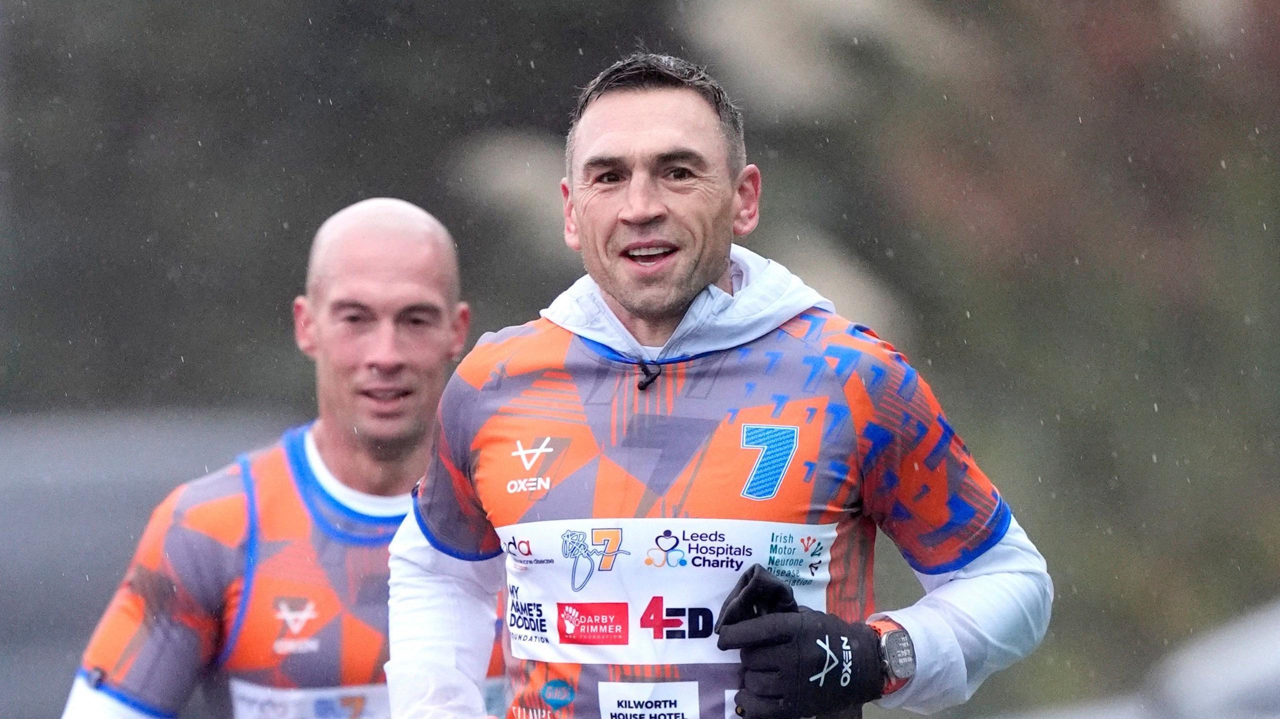 Kevin Sinfield running towards the camera, looking to the right. He has short, brown hair and is wearing a white, hooded jacket with an orange and blue T-shirt over the top. The T-shirt has multiple logos of charities on and a large, blue number seven. There is a man, who is bald, running behind him who is also wearing an orange and blue T-shirt. 