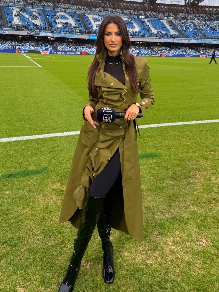A female sports reporter holding a DAZN microphone stands on a football pitch, wearing an olive trench coat, black top, leggings, and black patent leather boots.