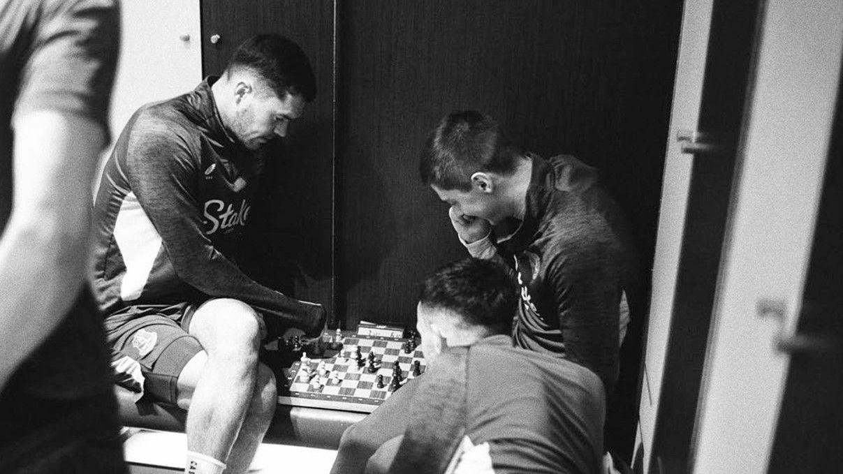 Two footballers wearing their Everton kits in the dressing room are playing chess. Two other players are watching on in this black and white image. 