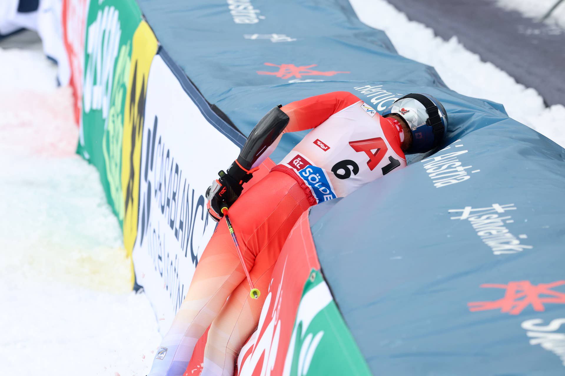 Ein Skifahrer im Rennanzug lehnt seitlich an der Bande nach einem Sturz im Zielbereich.
