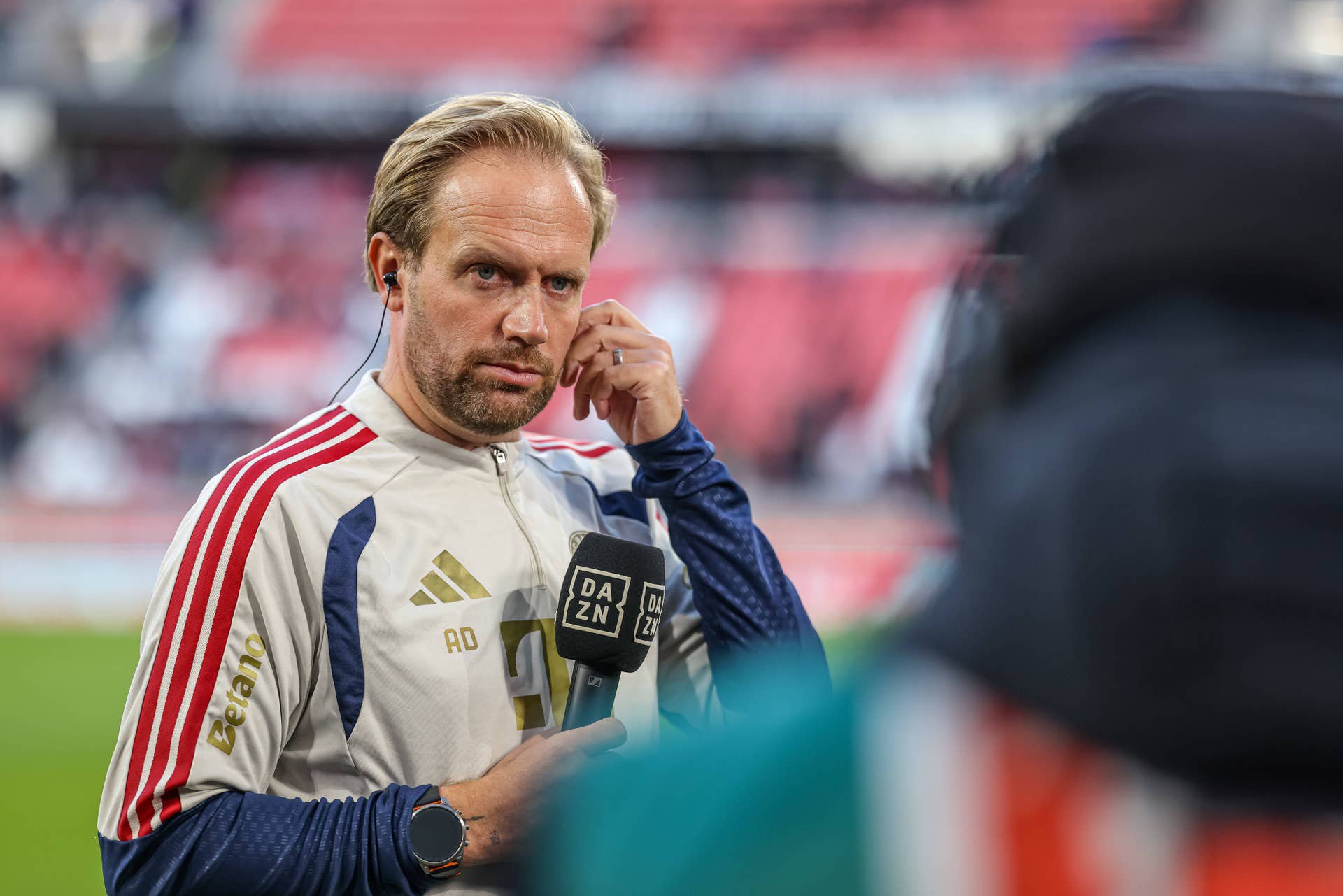 Ein Mann im Trainingsanzug spricht mit einem DAZN-Mikrofon auf einem Fußballfeld.