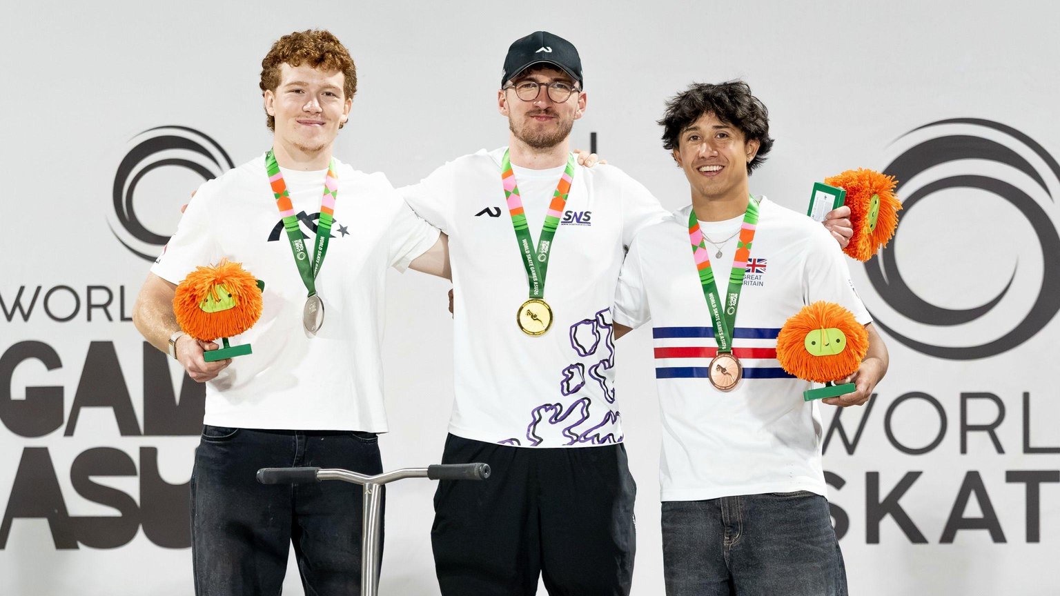 Three people standing on a podium, all with medals round their necks. Jayden Sharman, in the middle, is wearing a black cap, a white T-shirt and black trousers. He has glasses and a beard. The men next to him are dressed in white T-shirts and jeans and holding trophies.