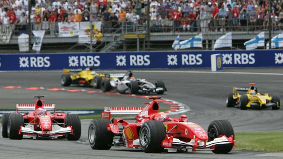 Ferrari lidera o GP dos EUA de F1 2005, com carros vermelhos na frente, Renault amarela ao fundo e público nas arquibancadas.