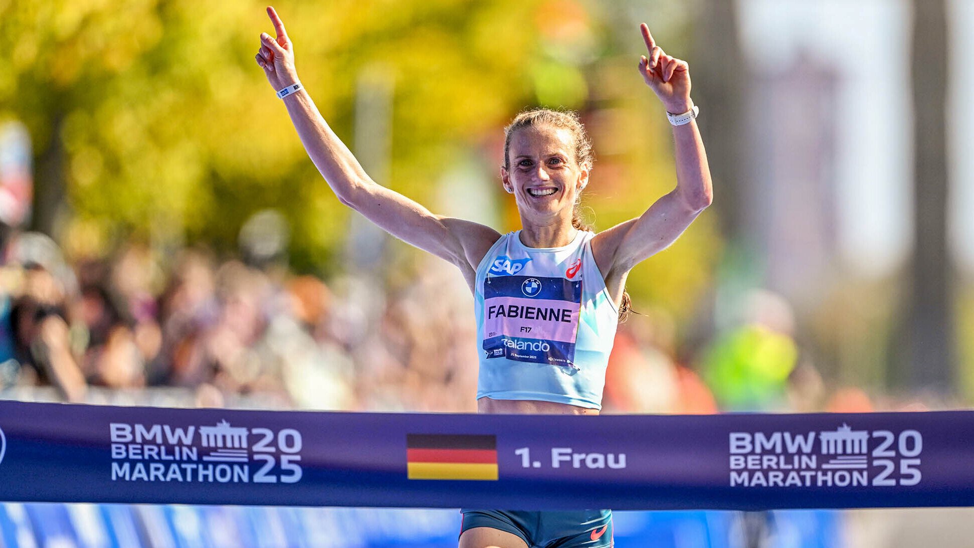 Fabienne Königstein läuft beim Berlin-Marathon 2025 mit erhobenen Armen jubelnd ins Ziel. Auf dem Zielband stehen eine Deutschlandfahne und „1. Frau“, sie war also die schnellste deutsche Läuferin.