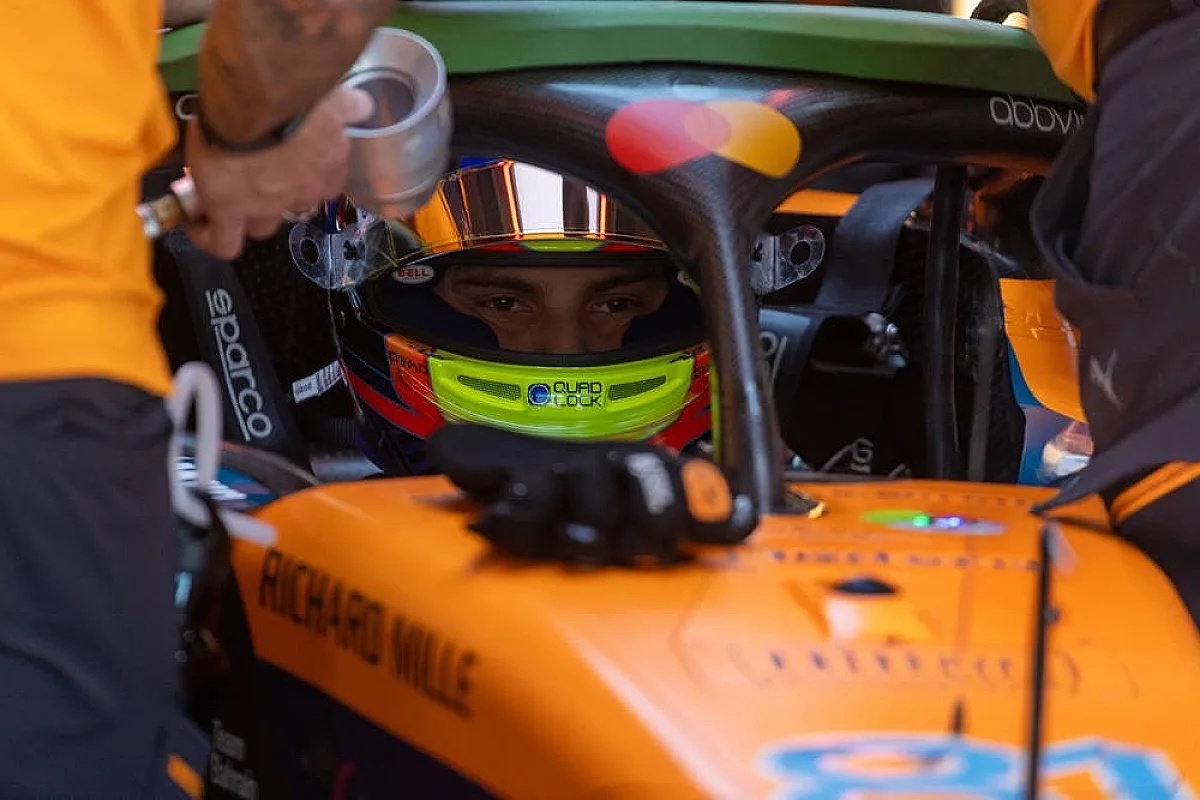 Driver in a McLaren F1 car in the pit garage, wearing a helmet with a green visor, with a McLaren mechanic in orange beside him