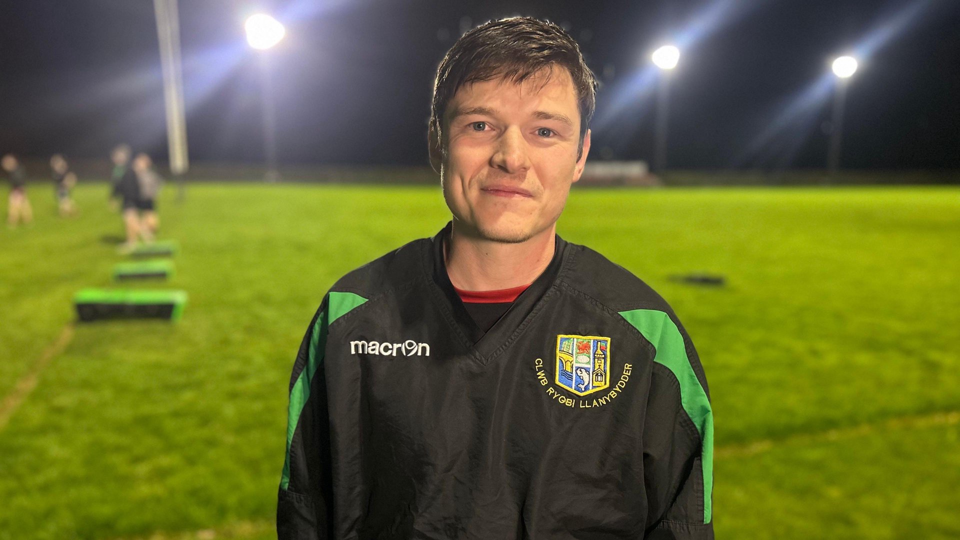 Dan Jones is photographed standing in front of a rugby pitch. He is wearing a black and green rugby jersey. Several men can be seen behind him wearing rugby kits. 