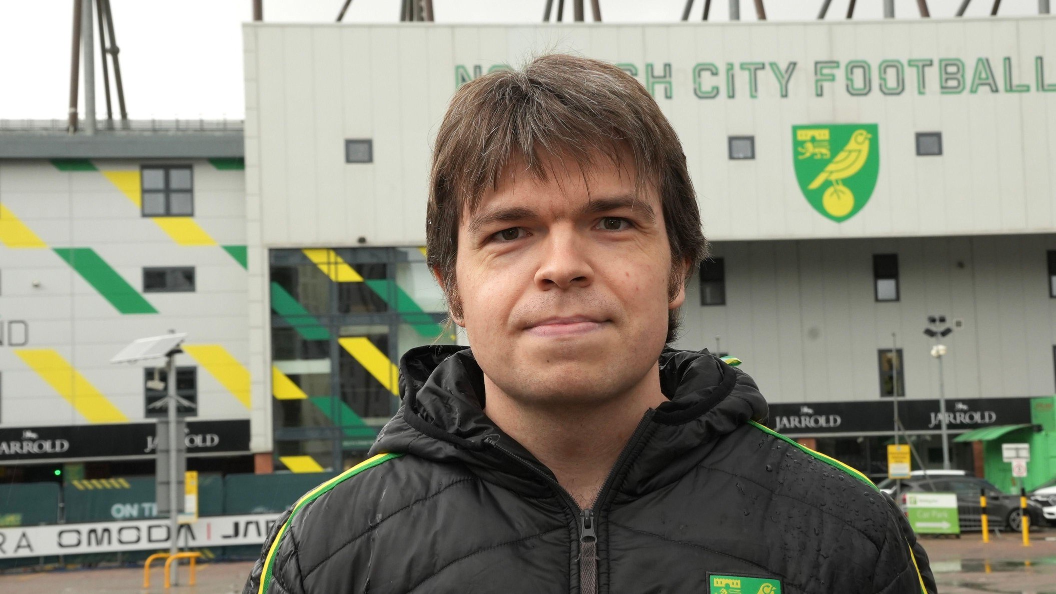 Tulsa outside the club's stadium at Carrow Road, which is behind him. Tulsa is wearing a black Norwich City jacket with the club's crest on the left of his chest. The words "Norwich City Football Club" are printed on the stadium roof behind.