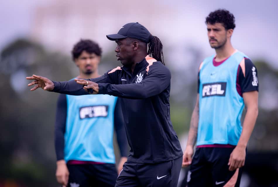 Treinador de futebol orienta treino com dois jogadores ao fundo, gesticulando, usando coletes azuis.