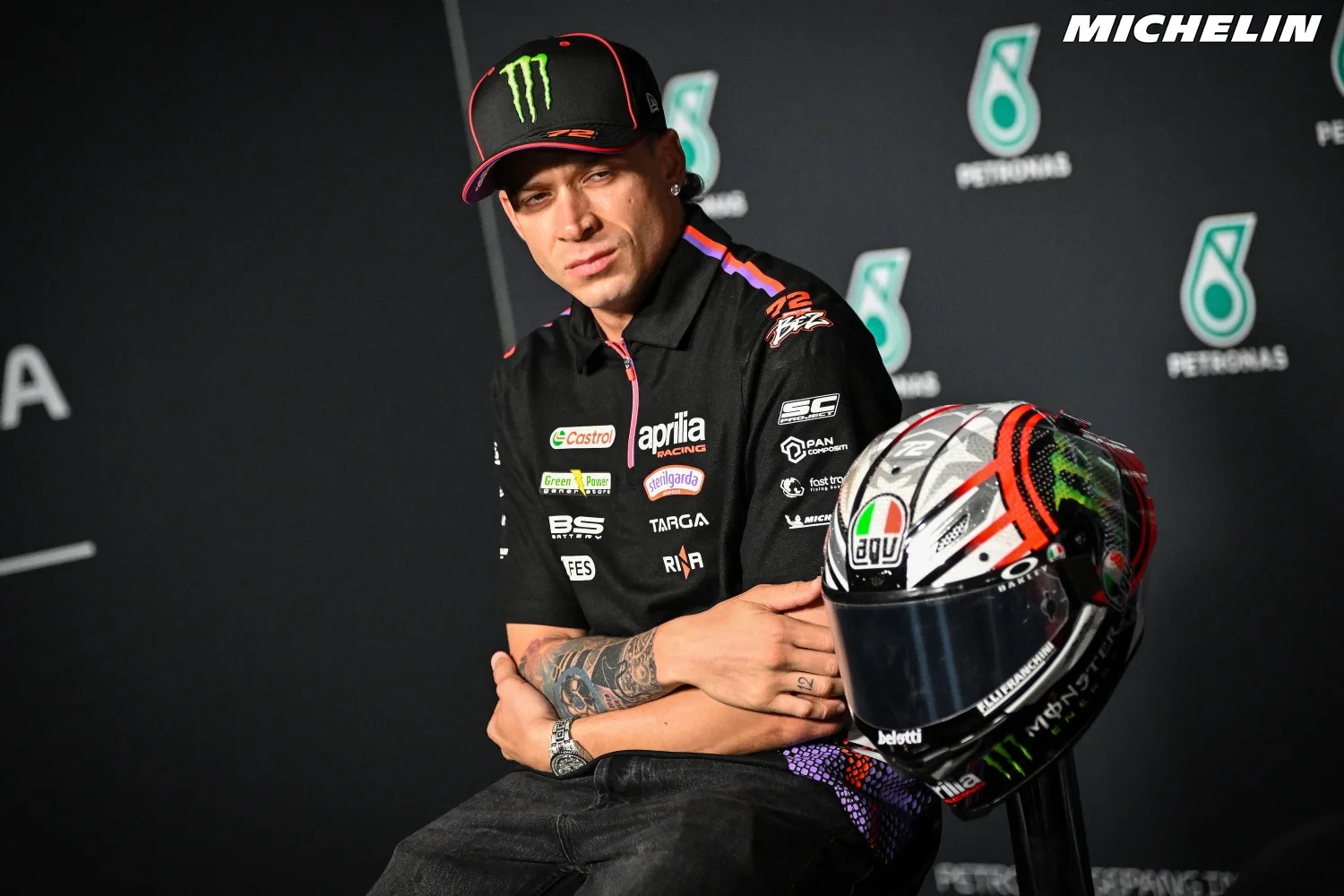 Rider in black Aprilia Racing shirt and cap with arms crossed beside a helmet at a MotoGP press conference, Petronas backdrop