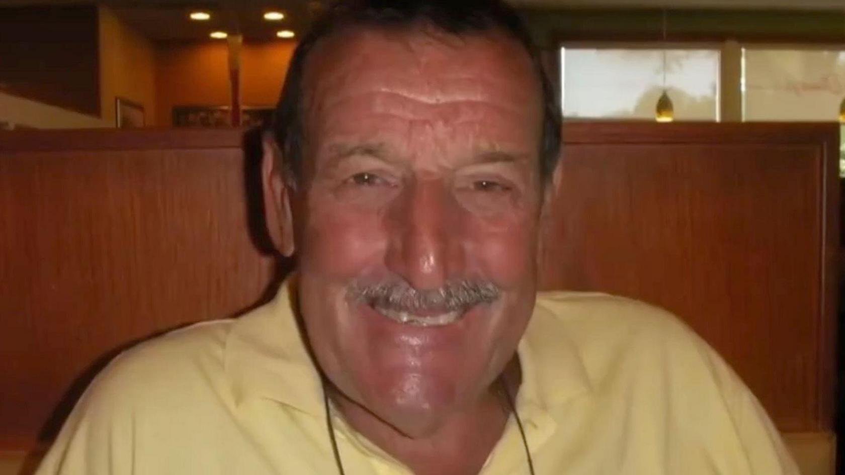 Close-up of Colin Flatt smiling. He has short dark hair and a moustache, which is greying. He is wearing a light yellow polo shirt. 