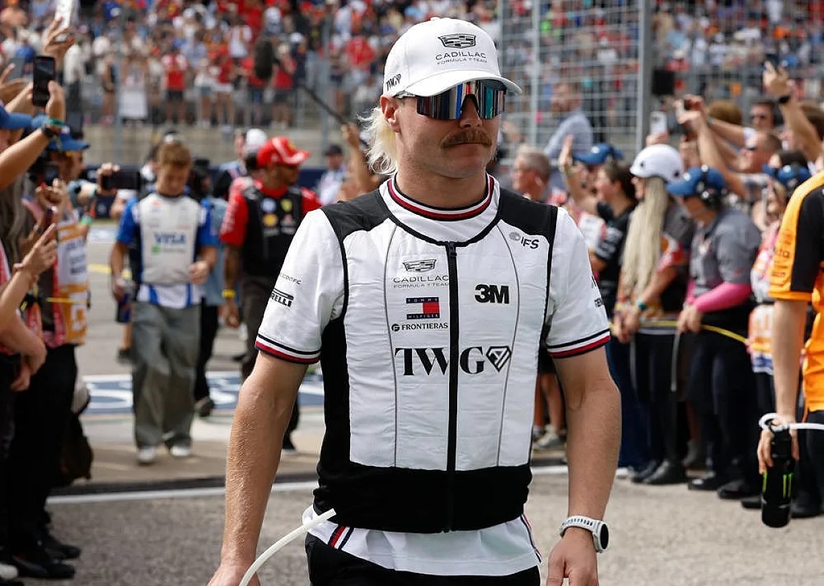 Driver in white Cadillac Formula 1 race suit and cap, sunglasses and moustache, walking through a fan-lined paddock area at Mexico City GP.