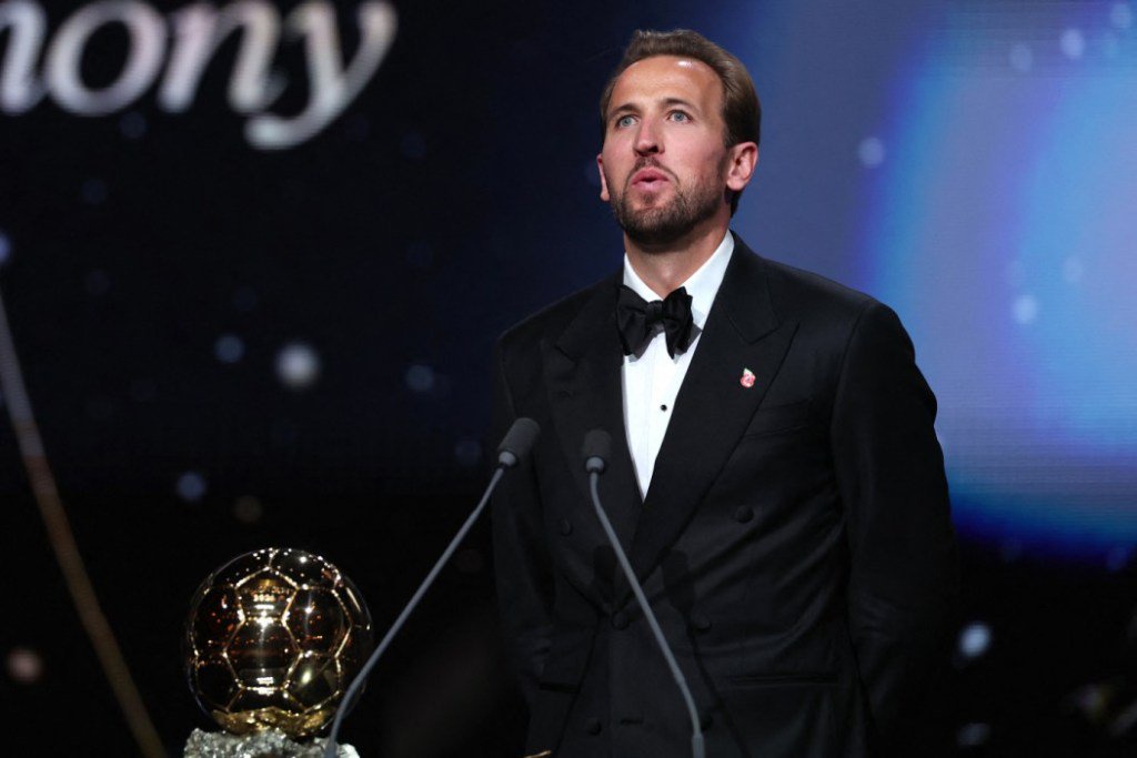 Harry Kane wearing a tuxedo at a podium with a Ballon d'Or trophy.