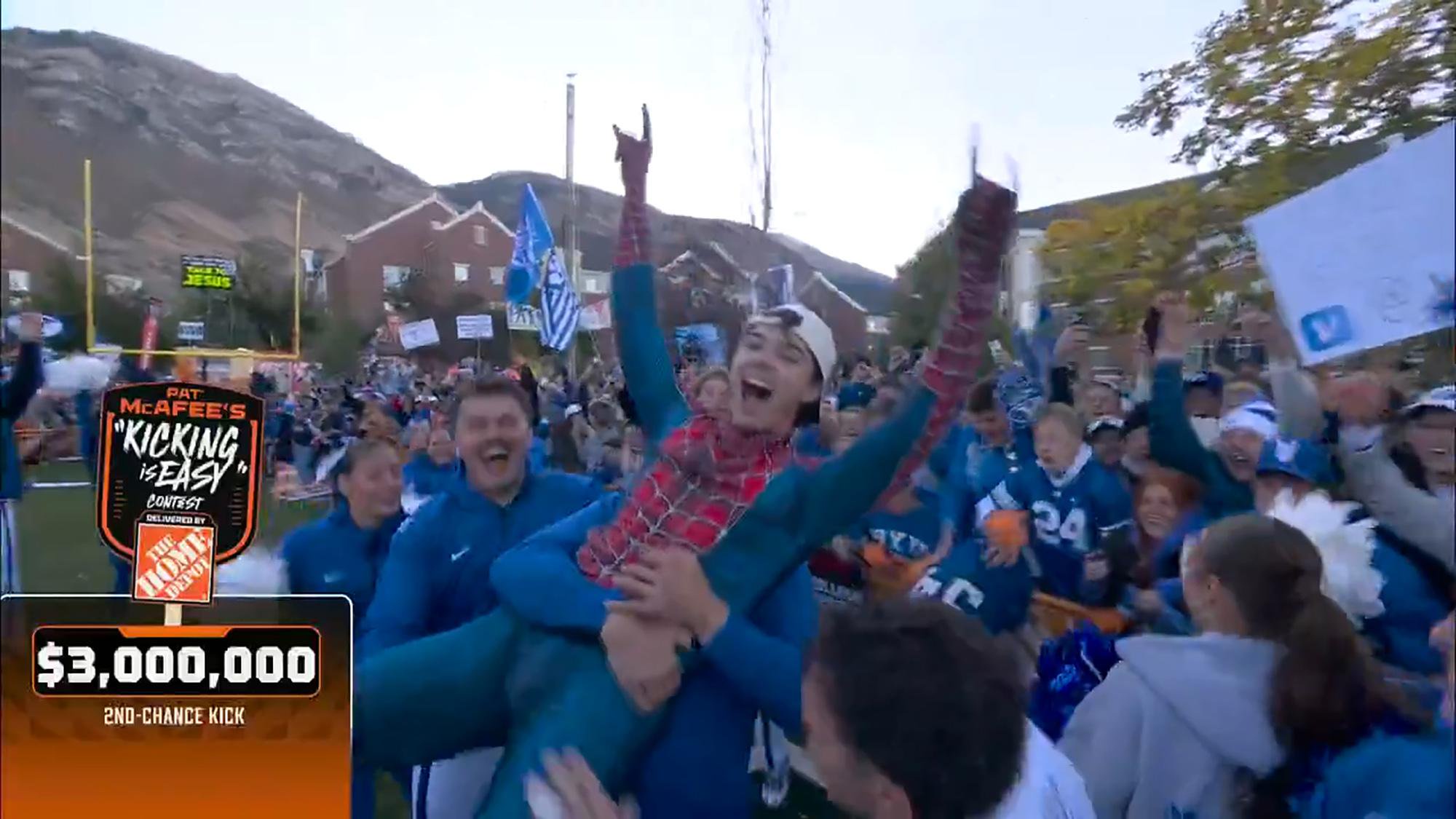 A man dressed as Spider-Man is lifted and carried joyfully by a cheering crowd on a college campus.
