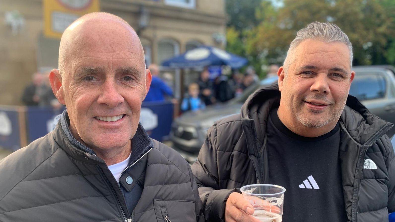 Phil Lockett (left) and Chris Helling (right) stand outside a pub or club venue before a match. Chris Helling holds a plastic cup of beer, while people and parked vehicles are visible in the blurred background.