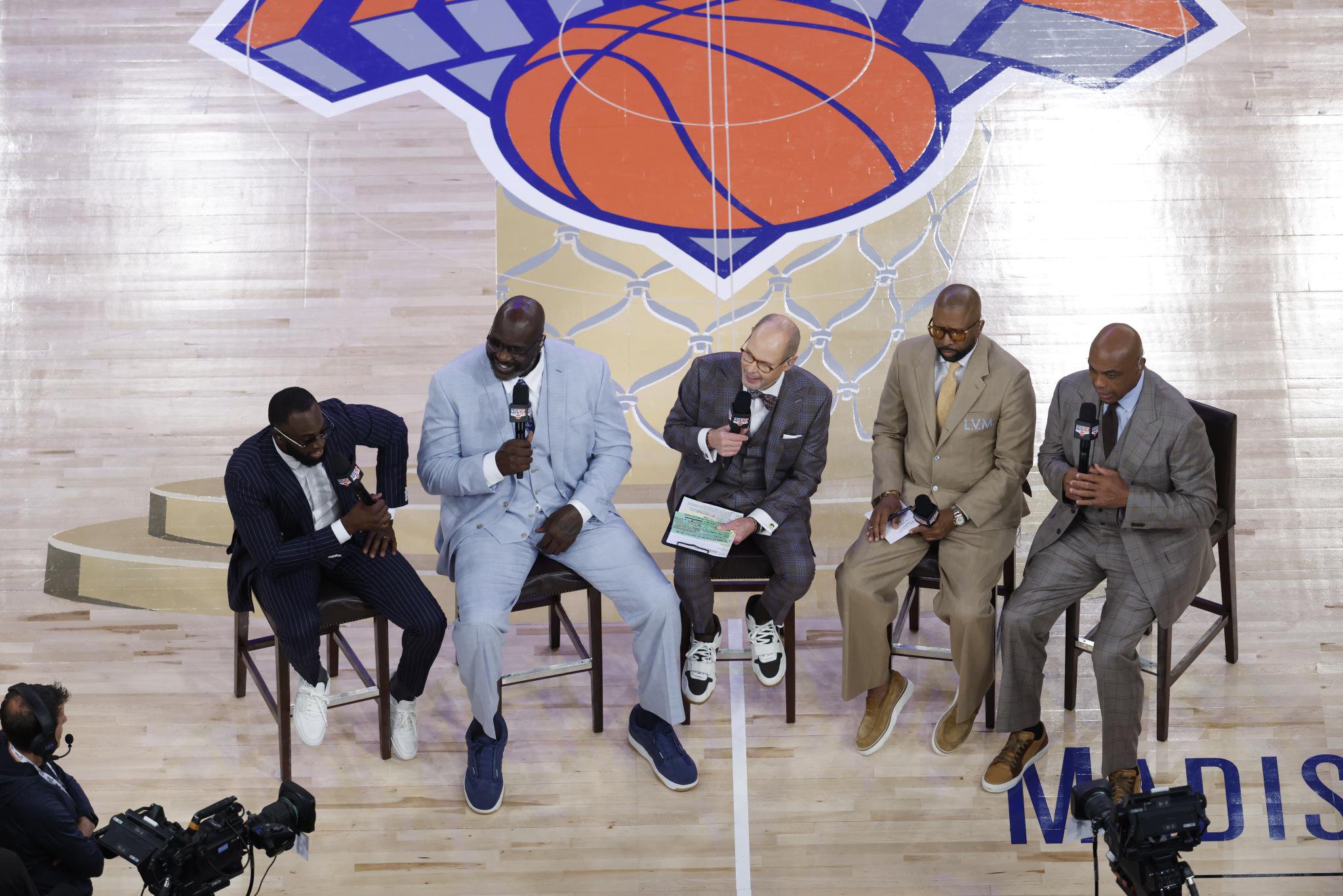 NBA broadcast analysts sitting on tall chairs on a basketball court, with a large New York Knicks logo painted on the floor behind them.