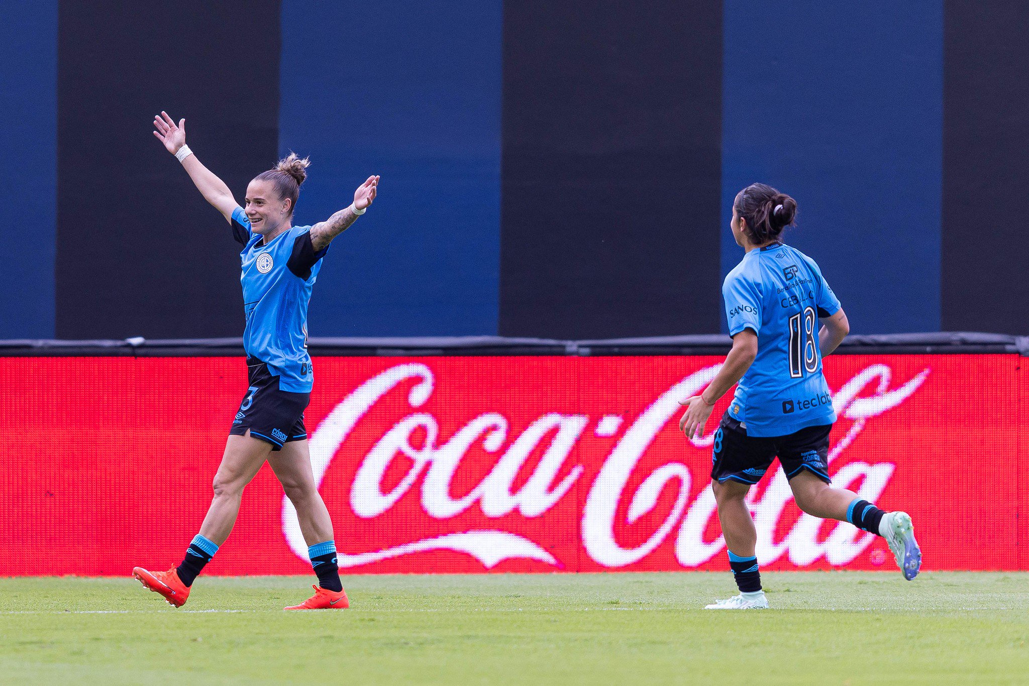 Duas jogadoras de futebol em campo, uma de costas com a camisa 18 e outra de frente com os braços erguidos em comemoração, ambas vestindo uniforme azul e preto. Ao fundo, um painel vermelho com o logo branco da Coca-Cola