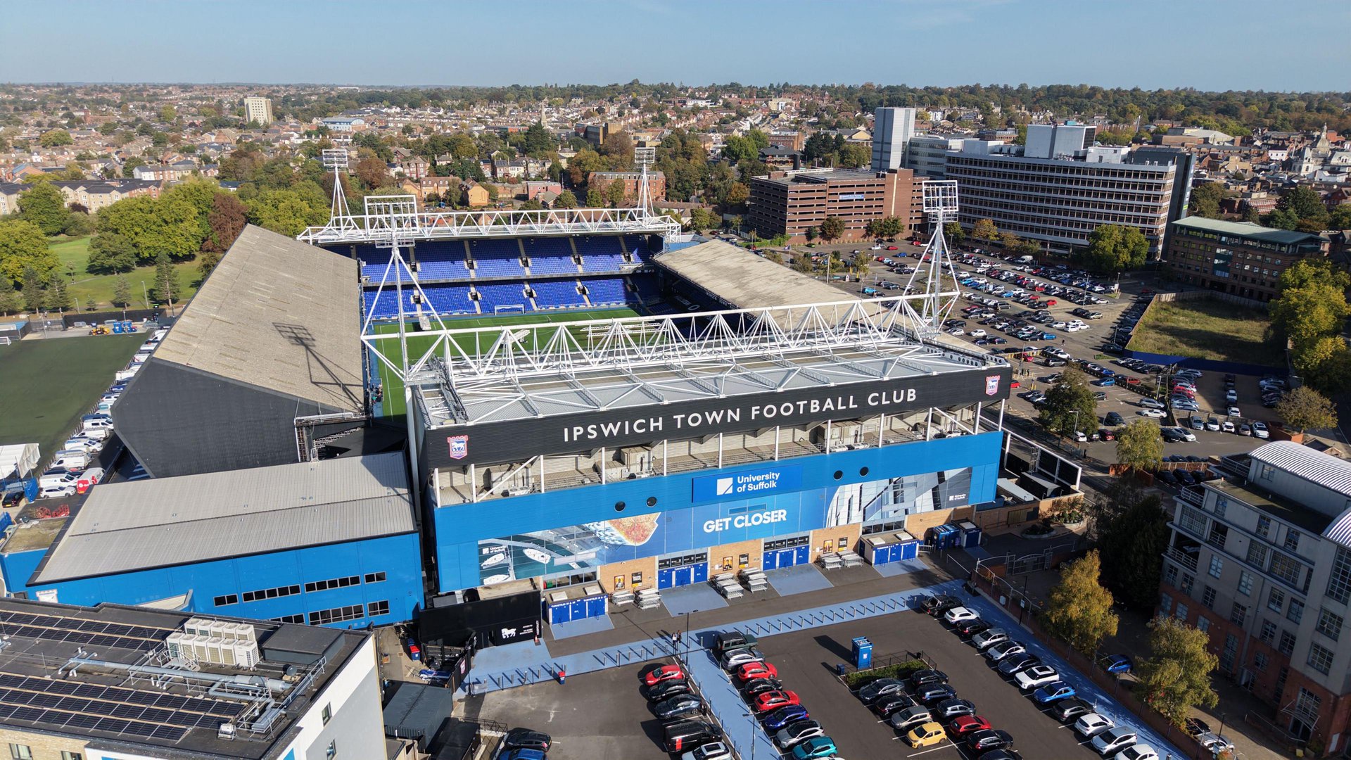 An aerial photograph showing Portman Road and the land surrounding, with a car park in the foreground and a view of the back of the Sir Alf Ramsey stand. It's a sunny day with blue sky.