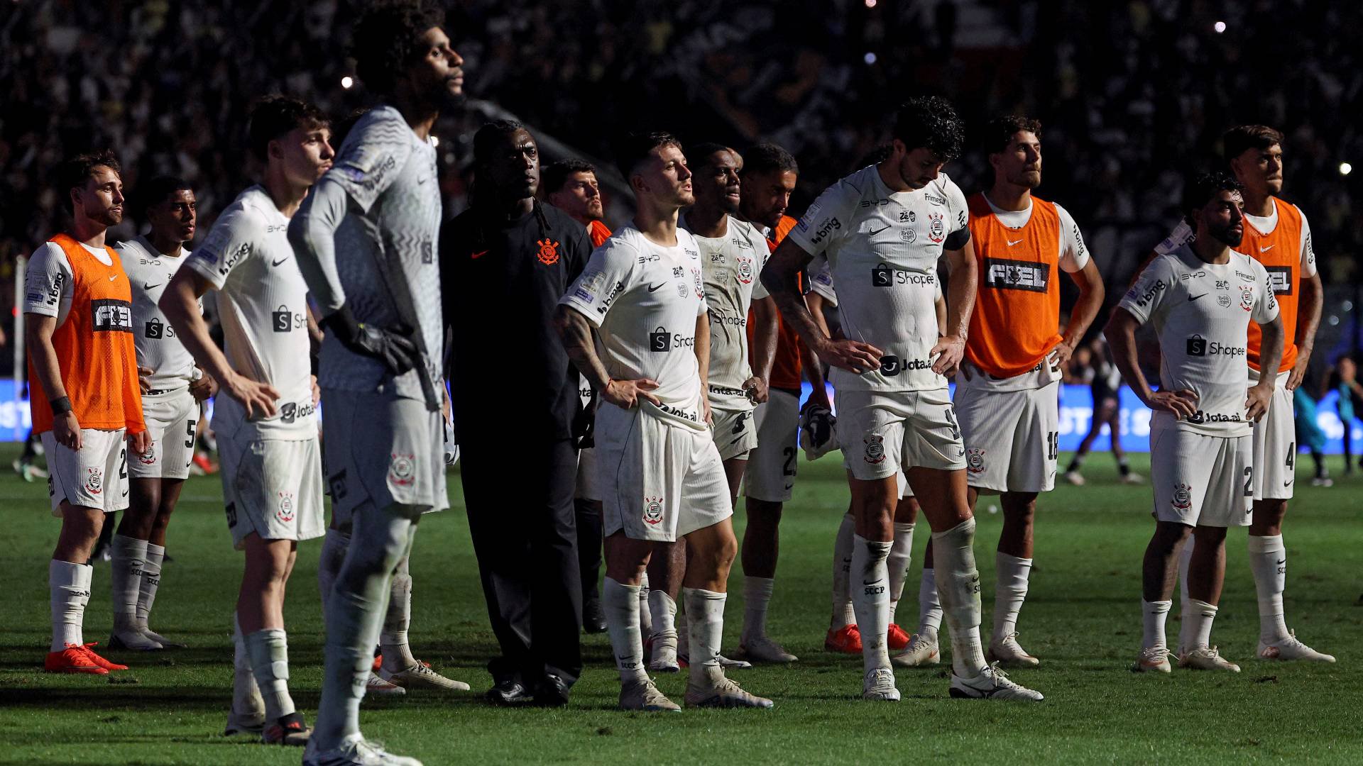 Jogadores de futebol do Corinthians, alguns com coletes laranjas, em pé no gramado verde de um estádio lotado à noite, com luzes brilhando ao fundo