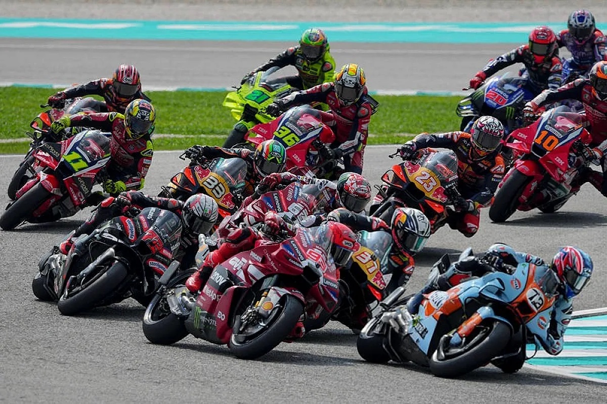 MotoGP riders racing in a tight pack at Sepang, including a red Ducati rider and a light blue bike ahead of the field, with the rain-affected track visible