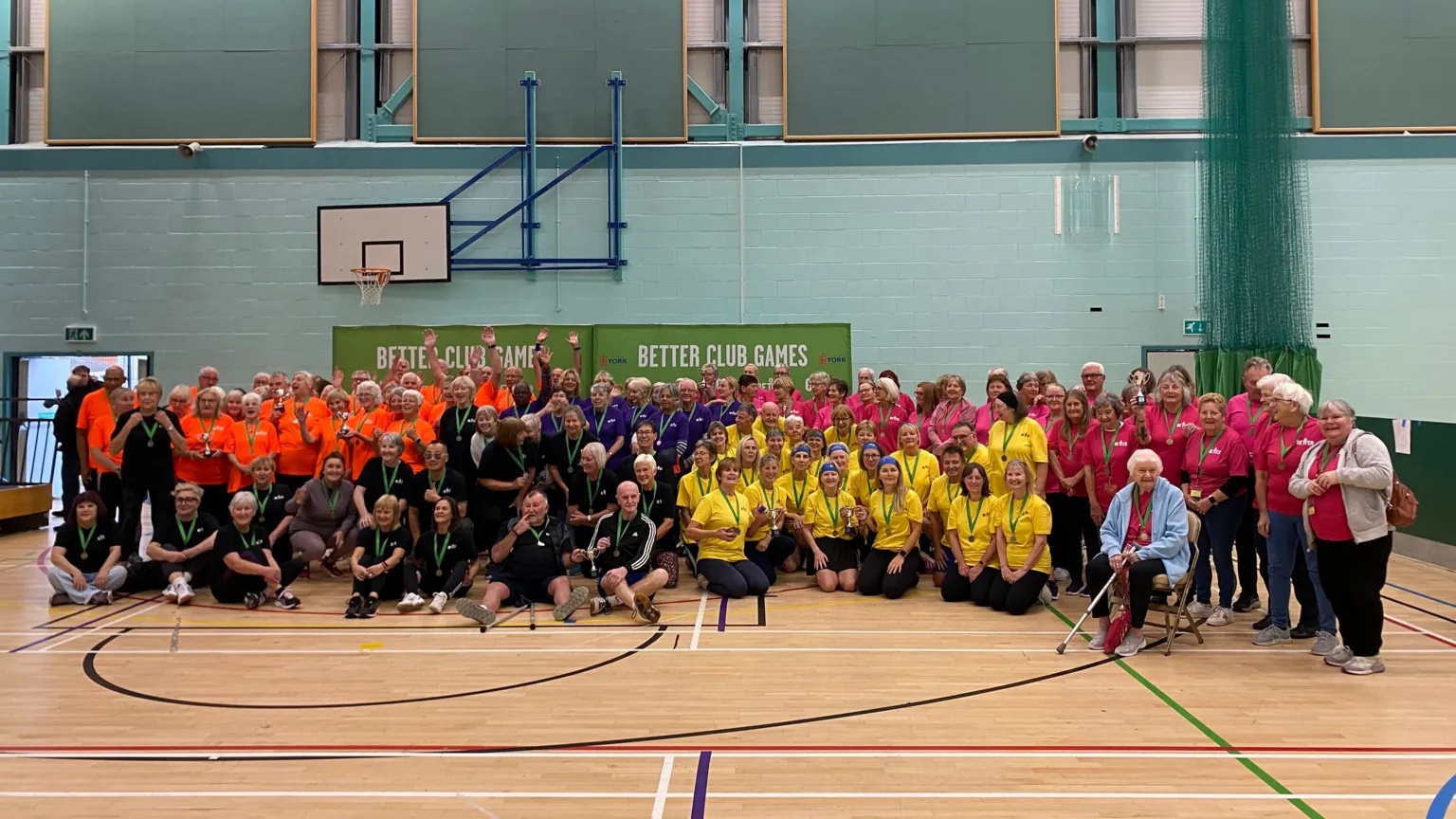 Large group photo in a sports hall showing dozens of participants wearing brightly coloured team shirts, including orange, pink and yellow. Many are wearing medals around their necks. There is a banner behind them that reads "Better Club Games". l.