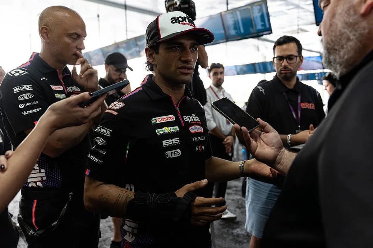 A rider in an Aprilia team shirt and cap talks to reporters in the MotoGP paddock, with microphones and phones held toward him.