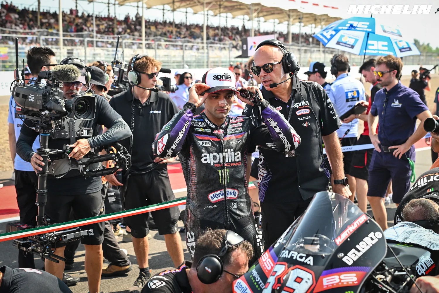 Jorge Martin on his Aprilia MotoGP bike in the pit lane, with cameras and crew around him and the Aprilia Racing logo visible.