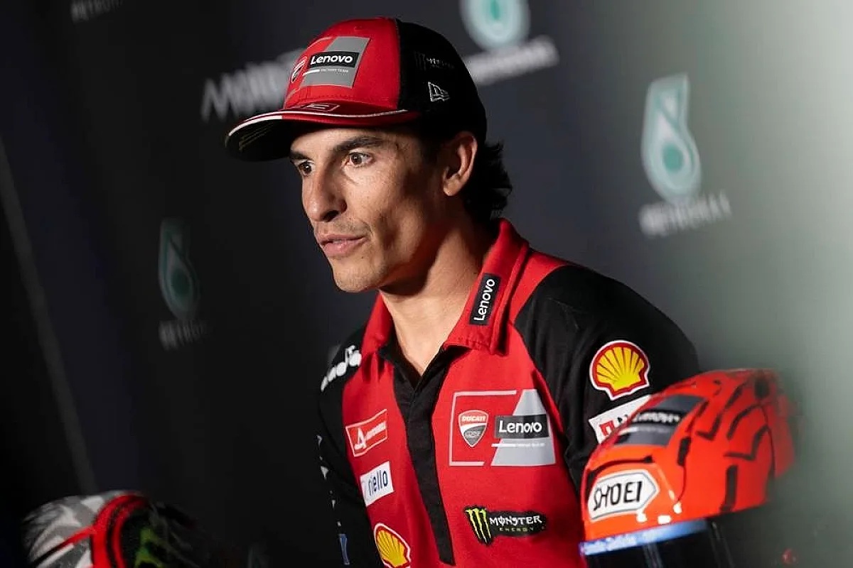 A rider in a red Ducati team cap and shirt with Lenovo and Shell logos, speaking at a MotoGP press conference, with an orange helmet in view.